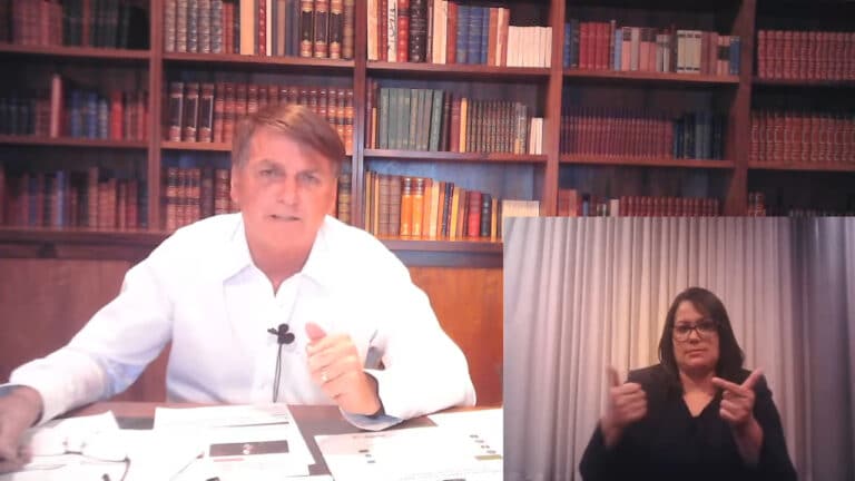 Imagem do presidente Jair Bolsonaro em transmissão nas redes sociais na última quinta-feira (25). Ele aparece com uma biblioteca ao fundo e sentado atrás de uma mesa com papeis. Uma intérprete de libras aparece no canto inferior direito da tela