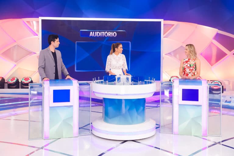 Imagem com foto do quadro Jogo das Três Pistas. Patricia Abravanel aparece ao centro, com os atores Velson D'Souza e Bianca Rinaldi em cada lado do palco