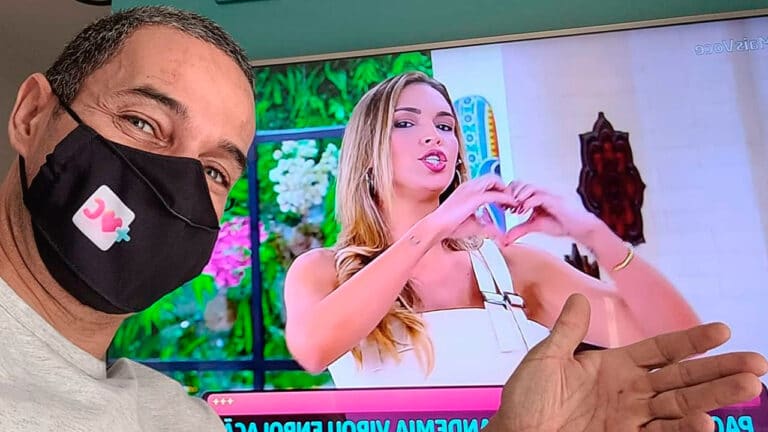 Imagem com foto de Fabricio Battaglini com Talitha Morete ao fundo na televisão