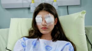 Imagem com foto da atriz Giovanna Grigio, intérprete de Mili, em cena da novela Chiquititas. Ela está com curativos nos olhos e deitada em uma cama de hospital