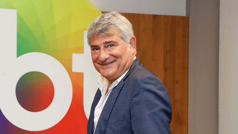 Cléber Machado na sala de reuniões de executivos do SBT sorrindo na frente do logotipo da emissora