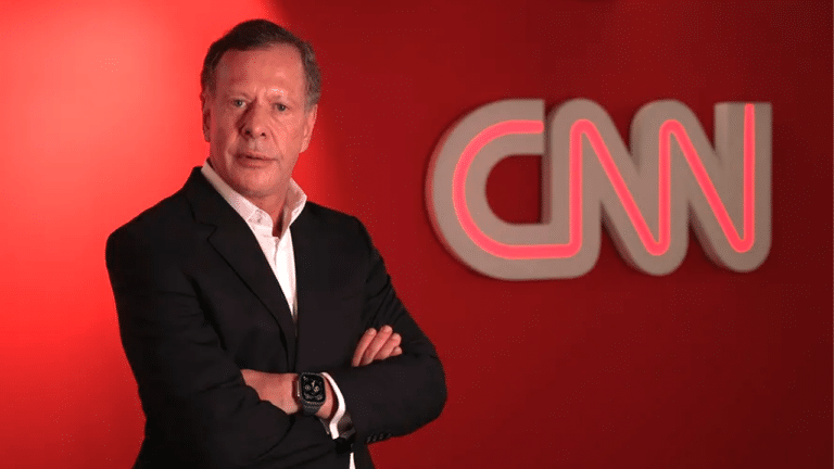 João Camargo, presidente da CNN Brasil, de braços cruzados em frente a uma parede vermelha com o logotipo da CNN