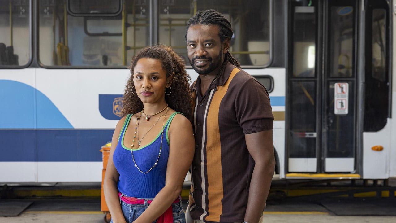 Madalena (Jéssica Ellen) e Jão (Fabrício Boliveira) sorridentes com um ônibus ao fundo