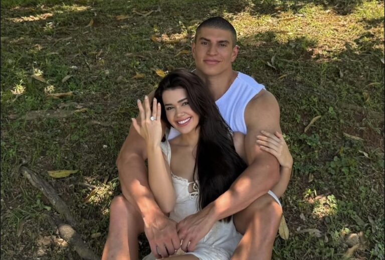 A imagem mostra a influenciadora Franciny Ehlke e seu noivo em um momento especial ao ar livre. Ela está sentada no colo dele, sorrindo e exibindo um anel de noivado reluzente na mão esquerda. Ele a abraça de forma protetora, vestindo uma regata branca que destaca seu porte físico