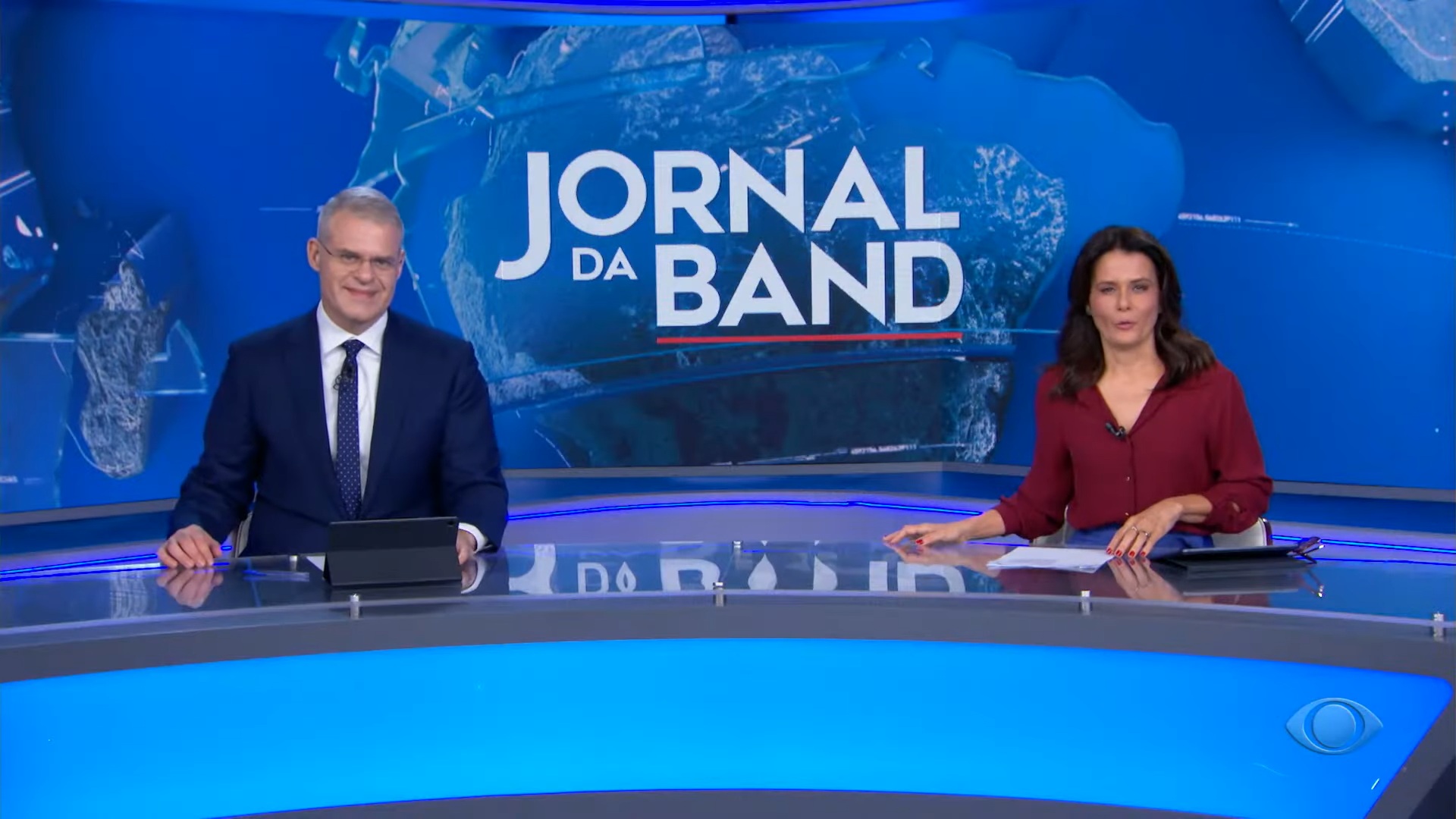 Foto dos jornalistas Eduardo Oinegue e Adriana Araújo na bancada do Jornal da Band