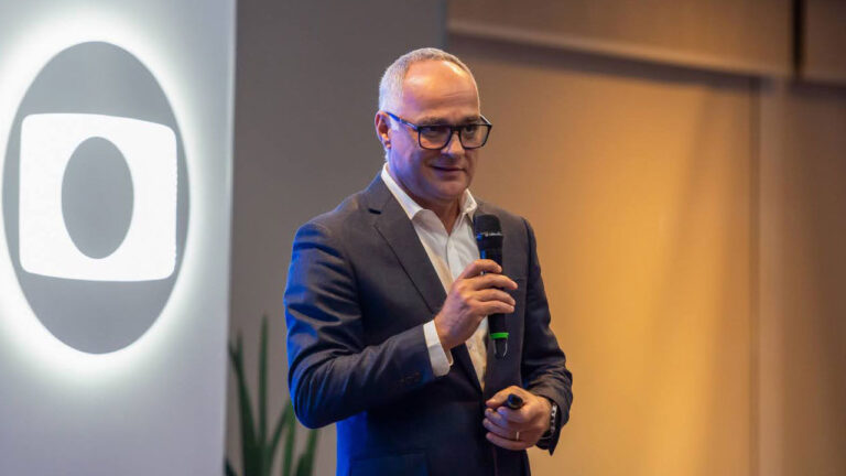 Amauri Soares, executivo da TV Globo, segurando um microfone durante apresentação, com o logotipo da emissora projetado ao fundo