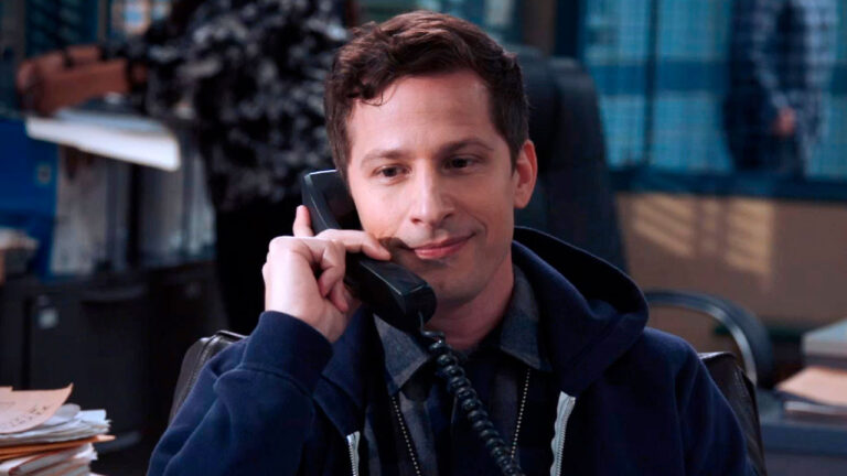 Andy Samberg como Jake Peralta em uma cena de Brooklyn Nine-Nine, segurando um telefone e sorrindo enquanto está sentado em sua mesa na delegacia