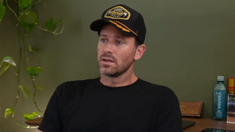 Armie Hammer sentado em um ambiente interno, vestindo camiseta preta e boné, com expressão séria e olhar direcionado para o lado