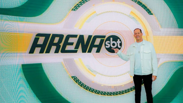 Benjamin Back posa sorrindo no estúdio do programa Arena SBT, apontando para o logo exibido em um painel digital com tons de verde e amarelo