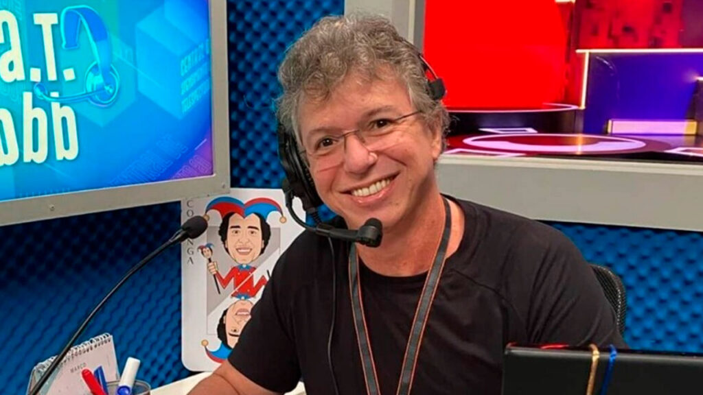 Boninho sorri enquanto usa um headset em um estúdio de gravação, com telas coloridas e itens do Big Brother Brasil ao fundo