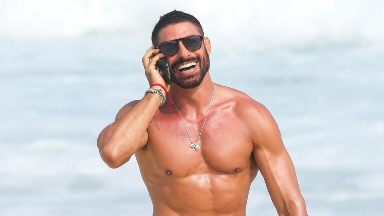 Cauã Reymond durante gravações do remake de Vale Tudo na praia da Barra da Tijuca, no Rio de Janeiro, sorrindo enquanto fala ao celular