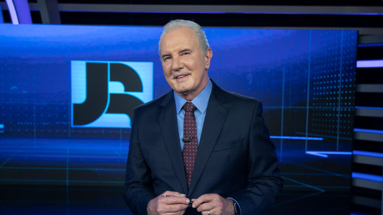 Celso Freitas no estúdio do Jornal da Record, vestindo terno escuro e gravata bordô, com o logotipo do telejornal ao fundo