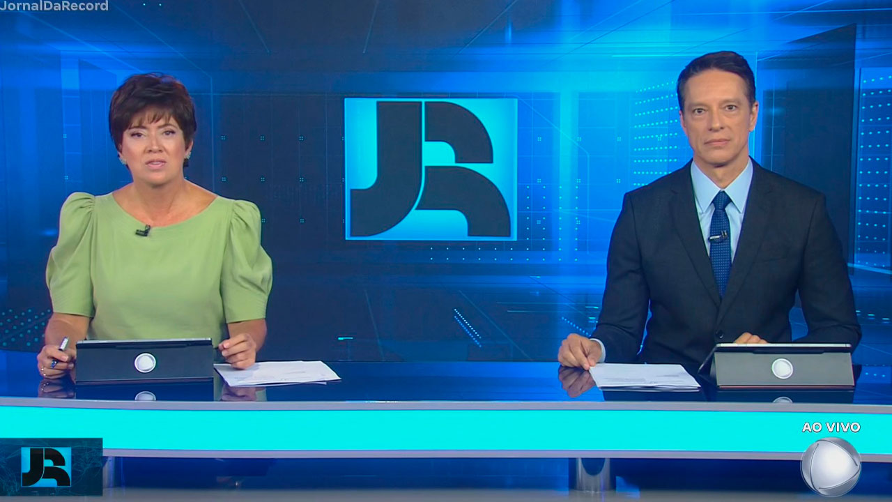 Christina Lemos e Sérgio Aguiar apresentando o Jornal da Record ao vivo, sentados na bancada com fundo azul e o logotipo do telejornal ao centro