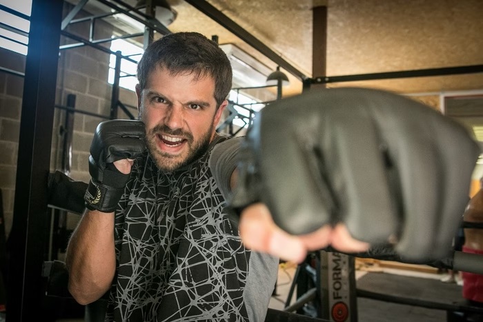 A imagem mostra Duda Nagle em um ambiente de treinamento de luta, possivelmente uma academia de artes marciais. O ator aparece vestindo uma camiseta esportiva preta com detalhes geométricos e luvas de proteção. Com uma expressão intensa e determinada, ele está na posição de ataque, lançando um soco em direção à câmera, o que cria um efeito dinâmico na imagem.