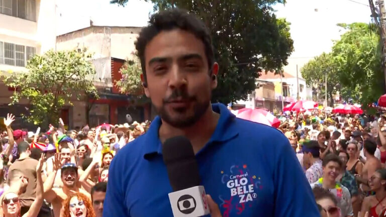 Fabrício Lobel, repórter da Globo, veste camisa azul com a logo do Carnaval Globeleza e segura um microfone enquanto cobre um bloco de rua lotado, com foliões animados ao fundo