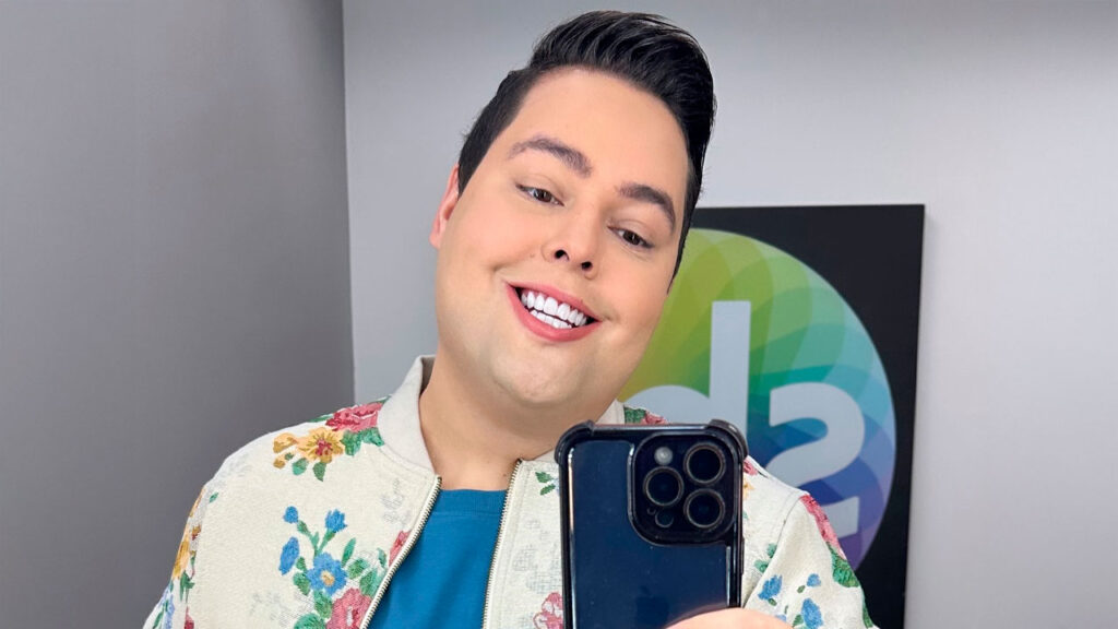 Fofoquito tirando uma selfie sorridente em frente a um painel com o logotipo do SBT