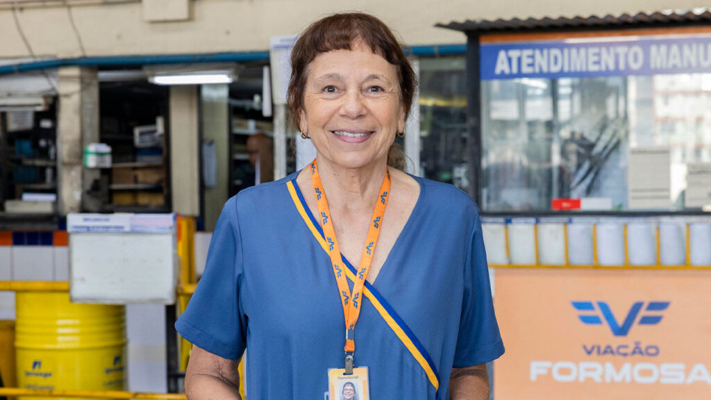 Iara Jamra sorrindo enquanto usa uniforme azul e crachá da Viação Formosa em um ambiente de atendimento da empresa fictícia da novela Volta por Cima