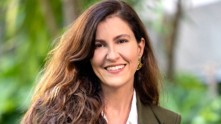 Leonora Bardini, diretora da Globo, sorrindo para a câmera, usando blazer verde e blusa branca, com cabelo solto e fundo desfocado com plantas verdes