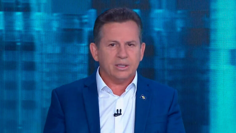 Mauro Mendes, governador do Mato Grosso, durante entrevista no programa Direto ao Ponto, da Jovem Pan, usando terno azul e falando diante de um fundo digital azul