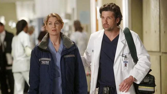 A imagem retrata uma cena icônica da série Grey’s Anatomy, mostrando os personagens Meredith Grey (Ellen Pompeo) e Derek Shepherd (Patrick Dempsey) em um corredor movimentado do hospital Grey Sloan Memorial.