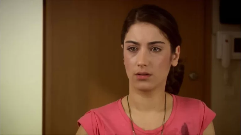 A imagem parece ser uma cena da novela O Segredo de Feriha (Adını Feriha Koydum), uma produção turca de grande sucesso. Na foto, a personagem principal, Feriha Yılmaz, interpretada por Hazal Kaya, aparece com uma expressão séria e preocupada. Ela veste uma blusa rosa e um colar fino, e seus cabelos estão presos em um rabo de cavalo baixo.