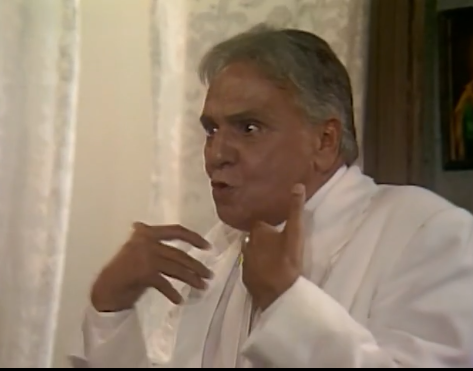 A imagem retrata o Pastor Hilário, personagem da icônica novela Tieta, exibida originalmente pela TV Globo em 1989. Ele aparece vestindo um terno branco, um de seus trajes característicos, e demonstra uma expressão intensa, com os olhos arregalados e a boca entreaberta, sugerindo que está no meio de um discurso inflamado ou reagindo a algo com indignação.