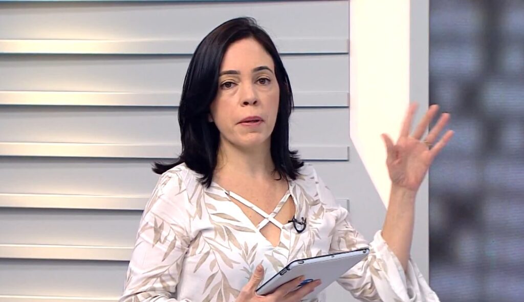 A imagem mostra a jornalista Patrícia Fiúza, conhecida por sua atuação na Globo Minas. Ela aparece apresentando um telejornal, segurando um tablet e gesticulando com a mão esquerda. Patrícia veste uma blusa branca com detalhes em bege e tem uma expressão séria e concentrada