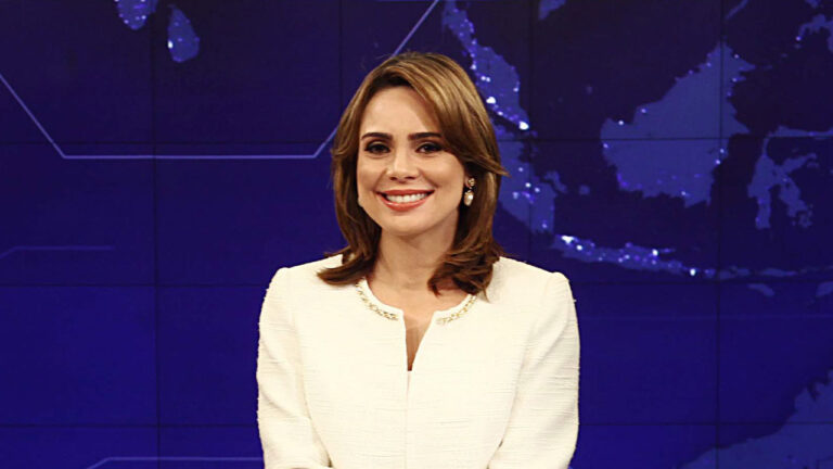 Rachel Sheherazade sorrindo na bancada do SBT Brasil, vestindo blazer branco, com fundo azul exibindo um mapa mundi iluminado