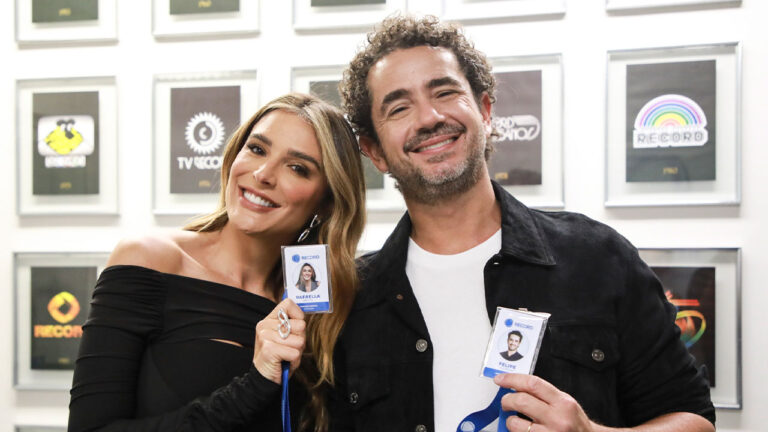 Rafa Brites e Felipe Andreoli sorrindo enquanto seguram crachás da Record, posando para foto em frente a uma parede com logotipos históricos da emissora