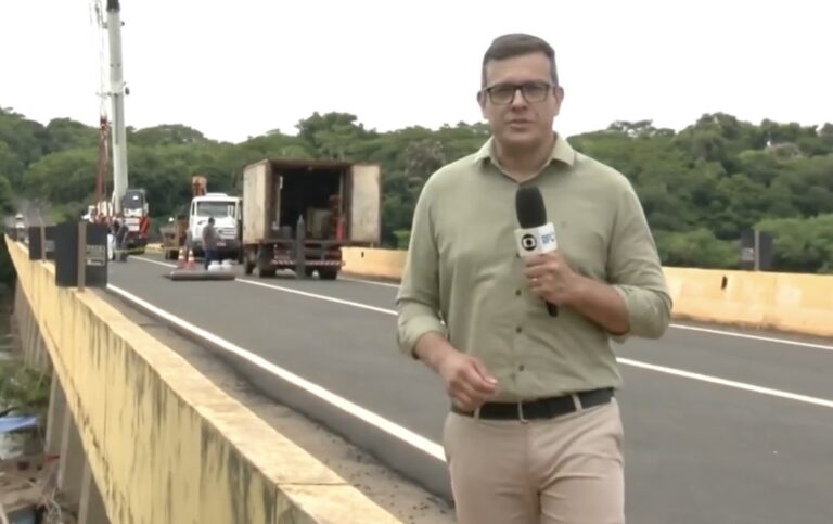 A imagem mostra o jornalista Rodrigo Grando durante uma reportagem em campo para a RPC, afiliada da Globo no Paraná. Ele aparece segurando um microfone com o logo da emissora, vestindo uma camisa social verde clara e calça bege, transmitindo um tom profissional e formal.