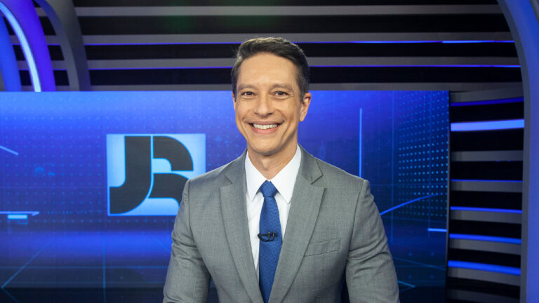 Sérgio Aguiar apresentando o Jornal da Record, sorrindo no estúdio com fundo digital azul e o logotipo do telejornal ao fundo