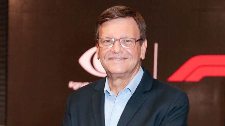 Sérgio Maurício sorrindo para a câmera em evento da Fórmula 1, usando terno preto e camisa azul com fundo escuro e logotipo da categoria