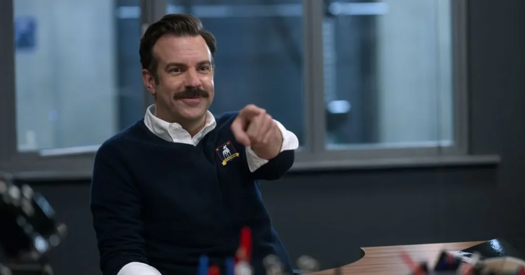 A imagem apresenta o personagem Ted Lasso, interpretado por Jason Sudeikis, em uma cena da aclamada série homônima da Apple TV+. Ele aparece sentado em um ambiente que remete a um escritório, vestindo um suéter azul-marinho com o brasão do AFC Richmond, equipe que ele comanda na série.