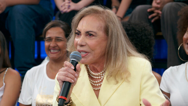 Arlete Salles de blazer amarelo e colar de pérolas fala ao microfone com a plateia do Altas Horas ao fundo