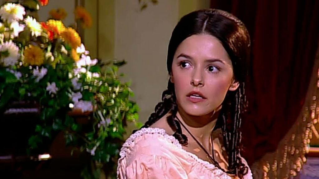 Bianca Rinaldi caracterizada como Isaura em cena da novela A Escrava Isaura com expressão de surpresa e flores ao fundo