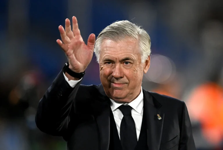Carlo Ancelotti, técnico do Real Madrid, aparece sorridente enquanto acena para o público. Usando terno preto, camisa branca e gravata escura, o treinador exibe expressão confiante e tranquila. A imagem, capturada em um estádio com iluminação noturna, transmite a postura elegante e vitoriosa que marca a carreira do comandante italiano