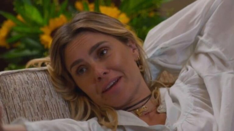 Carolina Dieckmann interpreta Leila em Vale Tudo. Na cena, a atriz aparece descontraída, recostada em um sofá, usando uma blusa branca de tecido leve. Com expressão serena e sorridente, ela exibe cabelos loiros soltos e usa colares delicados. Ao fundo, girassóis compõem o cenário, trazendo um toque de cor e leveza ao ambiente.