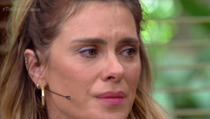 Carolina Dieckmann aparece emocionada durante participação no É de Casa, da Globo. Com lágrimas nos olhos, a atriz usa maquiagem suave, brincos dourados e microfone de lapela. O fundo desfocado com tons verdes sugere um ambiente externo ou decorado com plantas