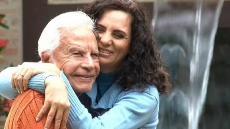 Cid Moreira aparece sorridente ao lado da esposa, Maria de Fátima Sampaio, em um momento de carinho e descontração. A jornalista o abraça por trás, com expressão afetuosa, enquanto ambos usam roupas de frio em tons suaves. O cenário ao fundo, com uma fonte de água e vegetação, transmite tranquilidade e intimidade.