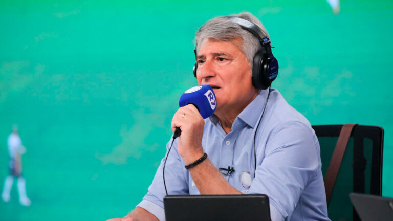 Cléber Machado usando fone de ouvido e narrando partida com microfone da Record frente a campo de futebol