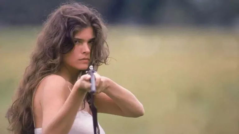 Cristiana Oliveira aparece caracterizada como Juma Marruá na primeira versão de Pantanal (1990). Com expressão séria e intensa, a atriz mira com uma espingarda, enquanto exibe cabelos longos e ondulados ao vento. Vestindo roupa simples, em meio a uma paisagem campestre, ela incorpora a força e a bravura de sua personagem icônica.
