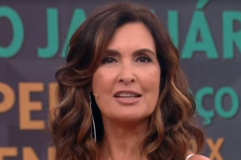 Fátima Bernardes aparece sorridente durante a apresentação de um programa de televisão. Com os cabelos castanhos ondulados e maquiagem leve, a jornalista usa um microfone de lapela discreto. Ao fundo, é possível ver um painel com letras grandes e coloridas, indicando um ambiente de estúdio vibrante e dinâmico.