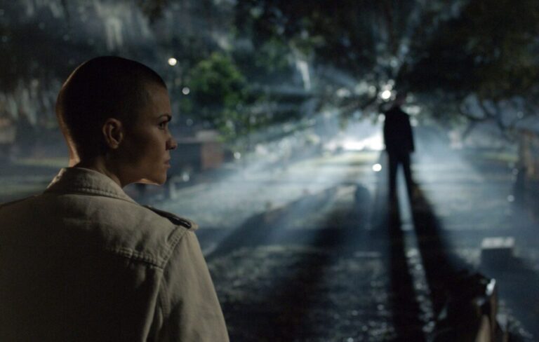 Cena do filme Conquista, que será exibido pela Record, mostra um momento de tensão em ambiente noturno. Em primeiro plano, uma personagem de cabeça raspada observa atentamente a figura de um homem à distância, envolto por feixes de luz entre as árvores. A atmosfera sombria e o jogo de luz e sombras criam um clima de suspense, típico das produções estreladas por Morgan Freeman.