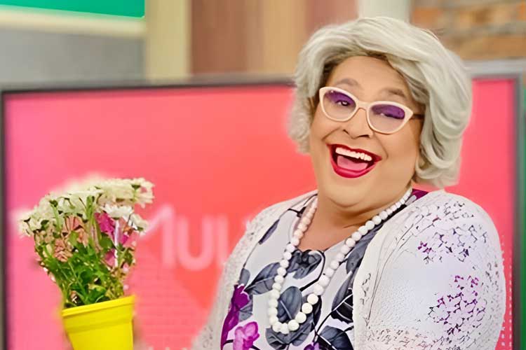 Guilherme Uzeda aparece caracterizado como Tia, sua personagem de sucesso, no estúdio do programa Mulheres, da TV Gazeta. Com peruca grisalha, óculos de armação clara, vestido florido e colar de pérolas, o artista segura um vaso de flores amarelo e sorri para a câmera em clima de bom humor