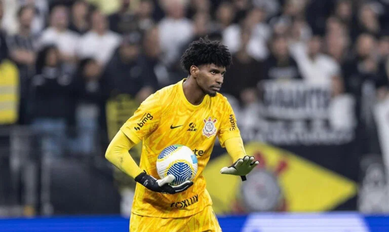 O goleiro Hugo Souza, do Corinthians, aparece em campo durante uma partida. Vestindo uniforme amarelo com detalhes pretos e segurando a bola com as duas mãos, o jogador demonstra concentração e postura de preparação. Ao fundo, torcedores e bandeiras do Corinthians preenchem as arquibancadas, reforçando o clima de tensão e expectativa no estádio.