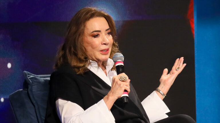 Iris Abravanel fala ao microfone sentada em poltrona durante evento, vestindo camisa branca e blazer preto