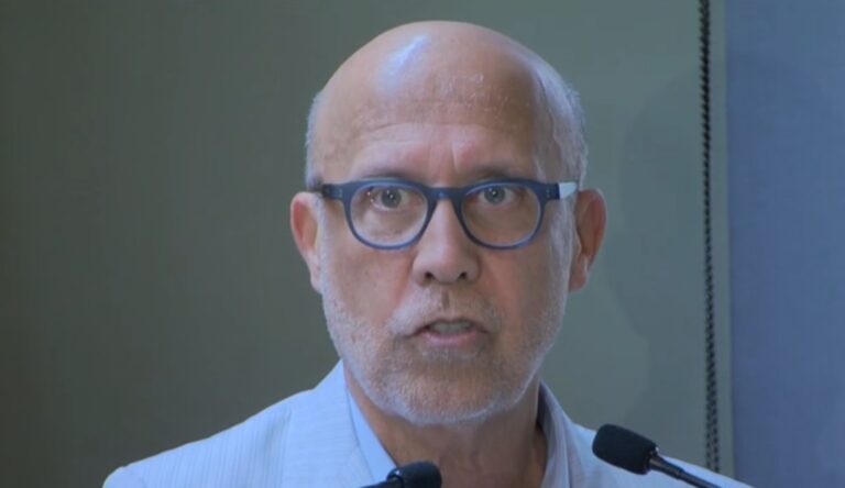 Jorge Nóbrega, executivo do Grupo Globo, aparece durante discurso em evento institucional. Na imagem, ele usa óculos de armação escura e traje claro, com microfone à frente, em ambiente formal