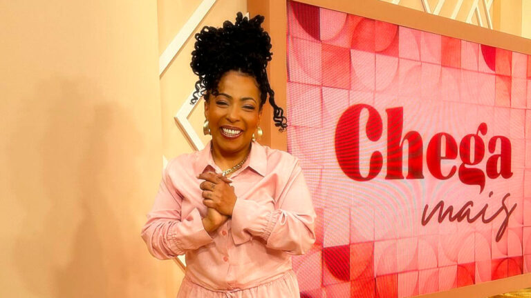 Juliana Oliveira sorrindo no cenário do programa Chega Mais, em frente ao telão com o nome da atração do SBT