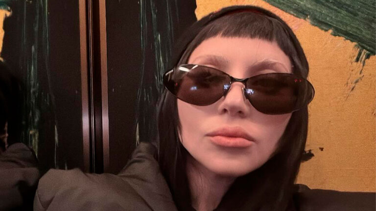 Lady Gaga com óculos escuros e cabelo preto liso, posando com expressão séria diante de fundo artístico