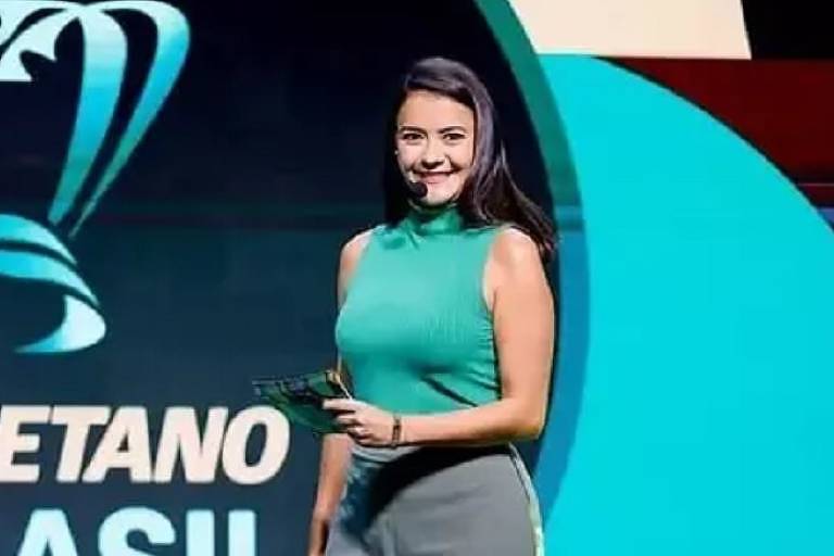 Lívia Torres surge sorridente durante a apresentação de um evento esportivo. Vestindo uma blusa verde sem mangas e calça cinza, a jornalista segura um cartão de apresentação e usa microfone de lapela. O fundo destaca o logotipo da competição, reforçando o clima dinâmico e profissional da transmissão.